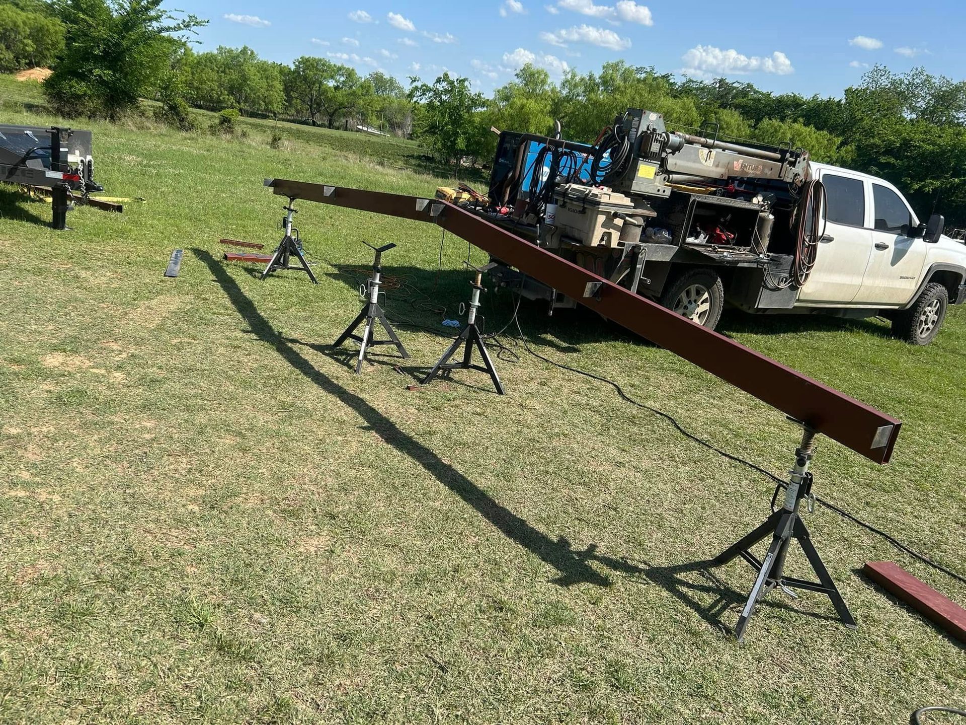 A long brown metal beam rests on three tripod stands in a grassy field next to a white service truck.
