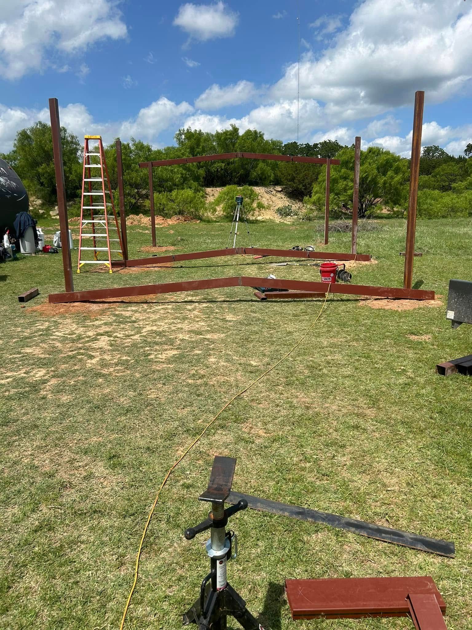 A metal pole structure under construction in a grassy field, featuring a tripod stand in the foreground.