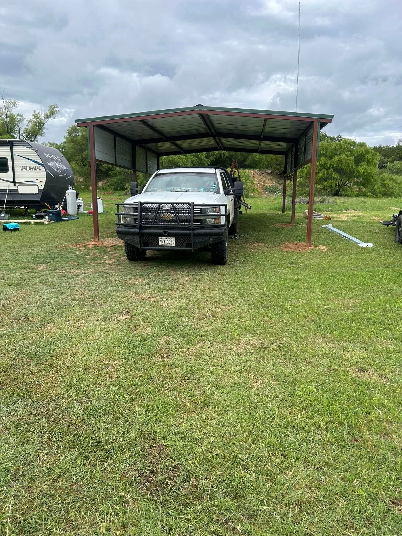 A white pickup truck with a heavy-duty bumper guard parked under a metal-roofed carport in a grassy field.