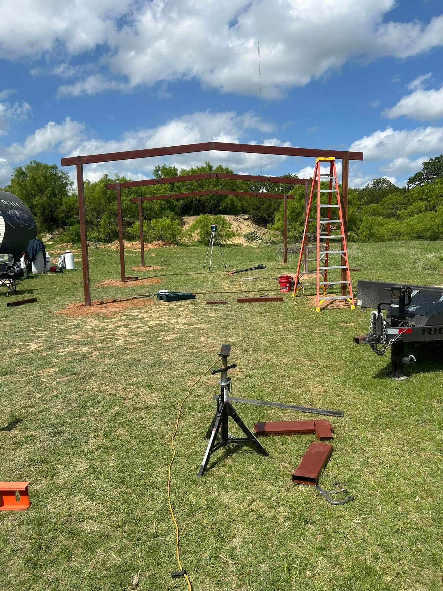 A partially constructed metal frame structure stands in a grassy field under a sunny sky, with a ladder and tripod nearby.