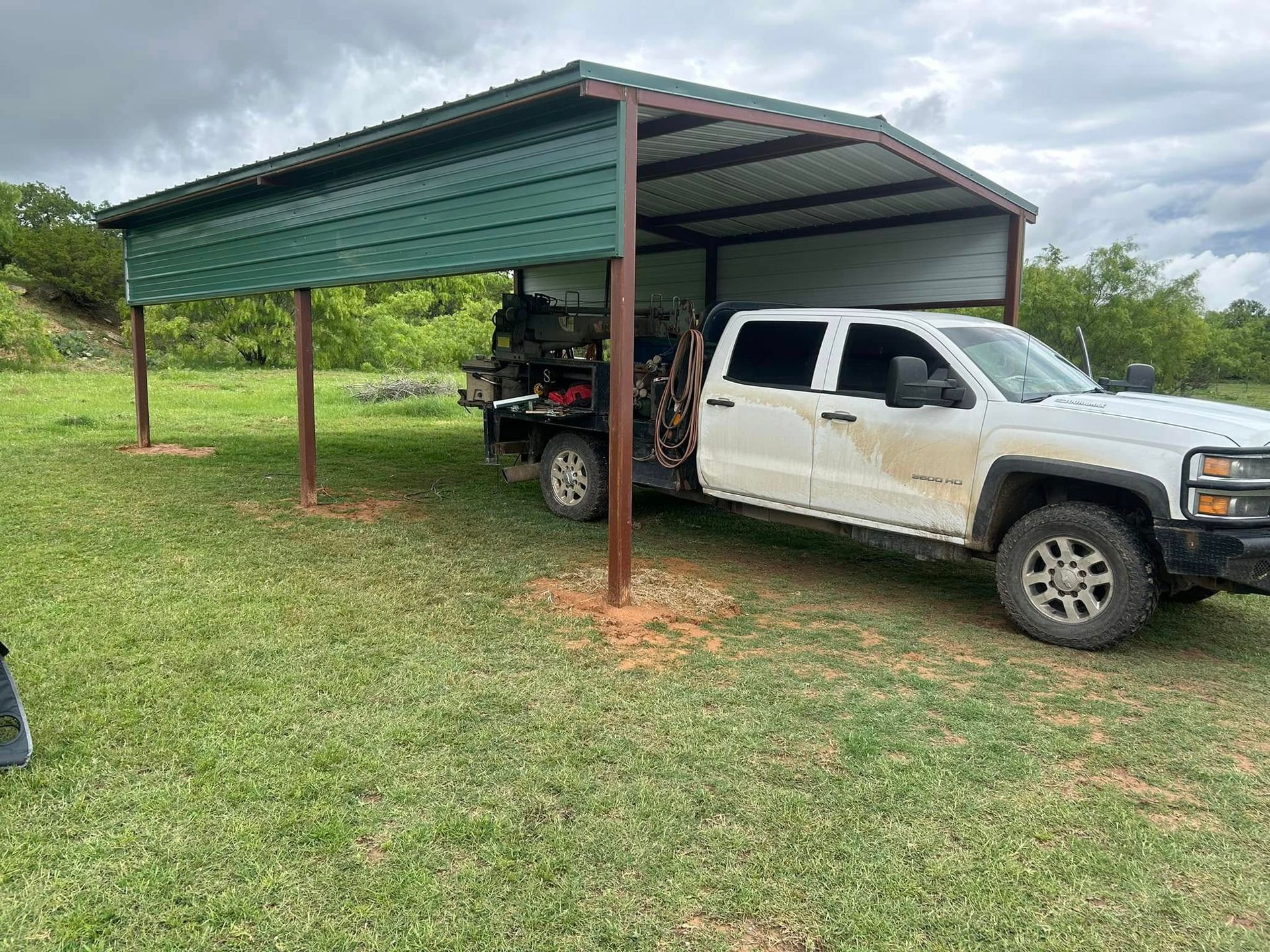 A white pickup truck parked under a metal-roofed green and silver carport in a grassy field.