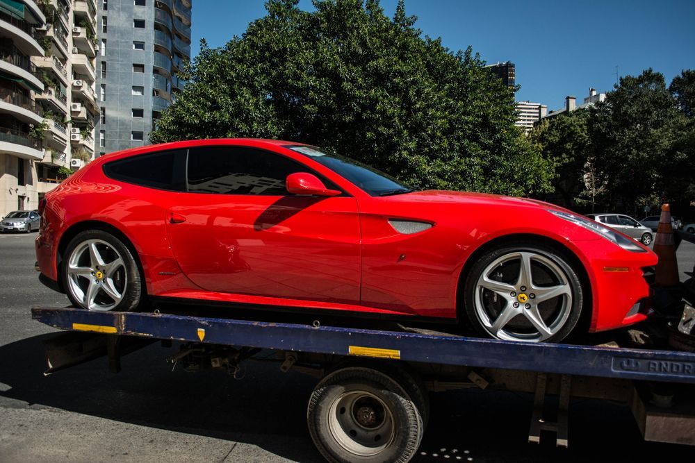 Red sports car on a tow truck in an urban street, with buildings and trees in the background