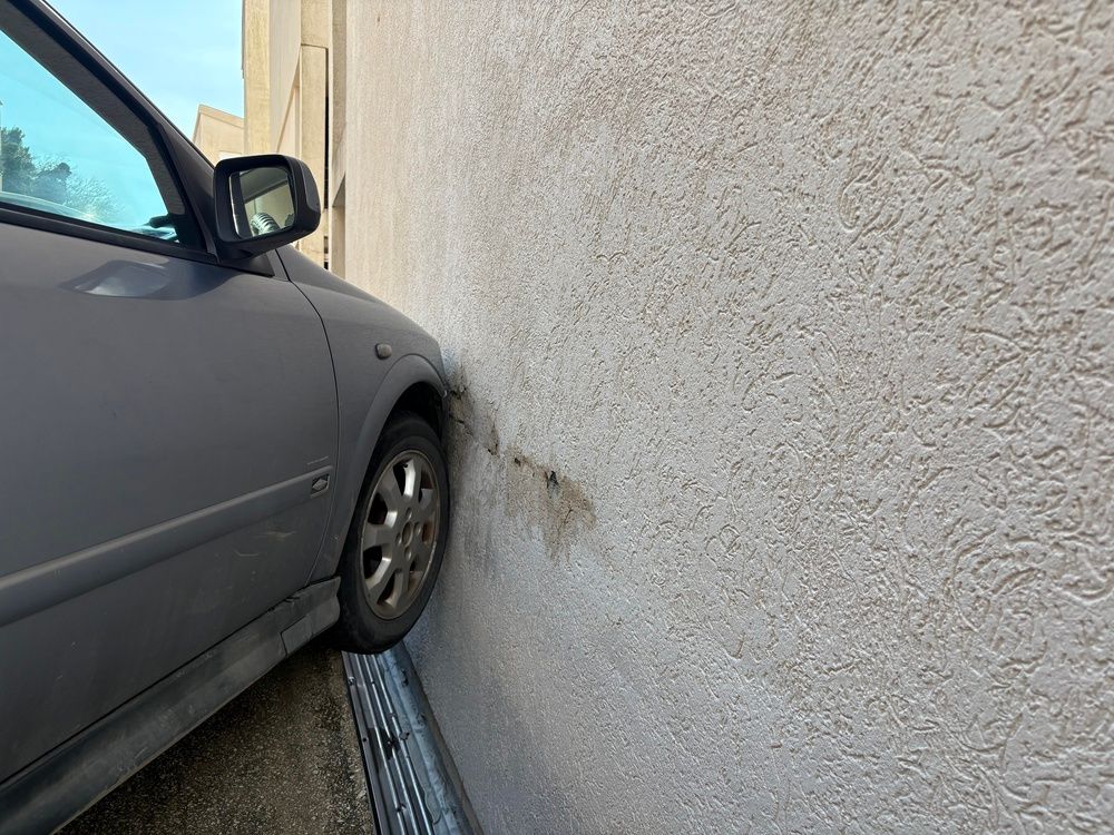 Gray car parked very close to a textured wall, with the front wheel nearly touching it.