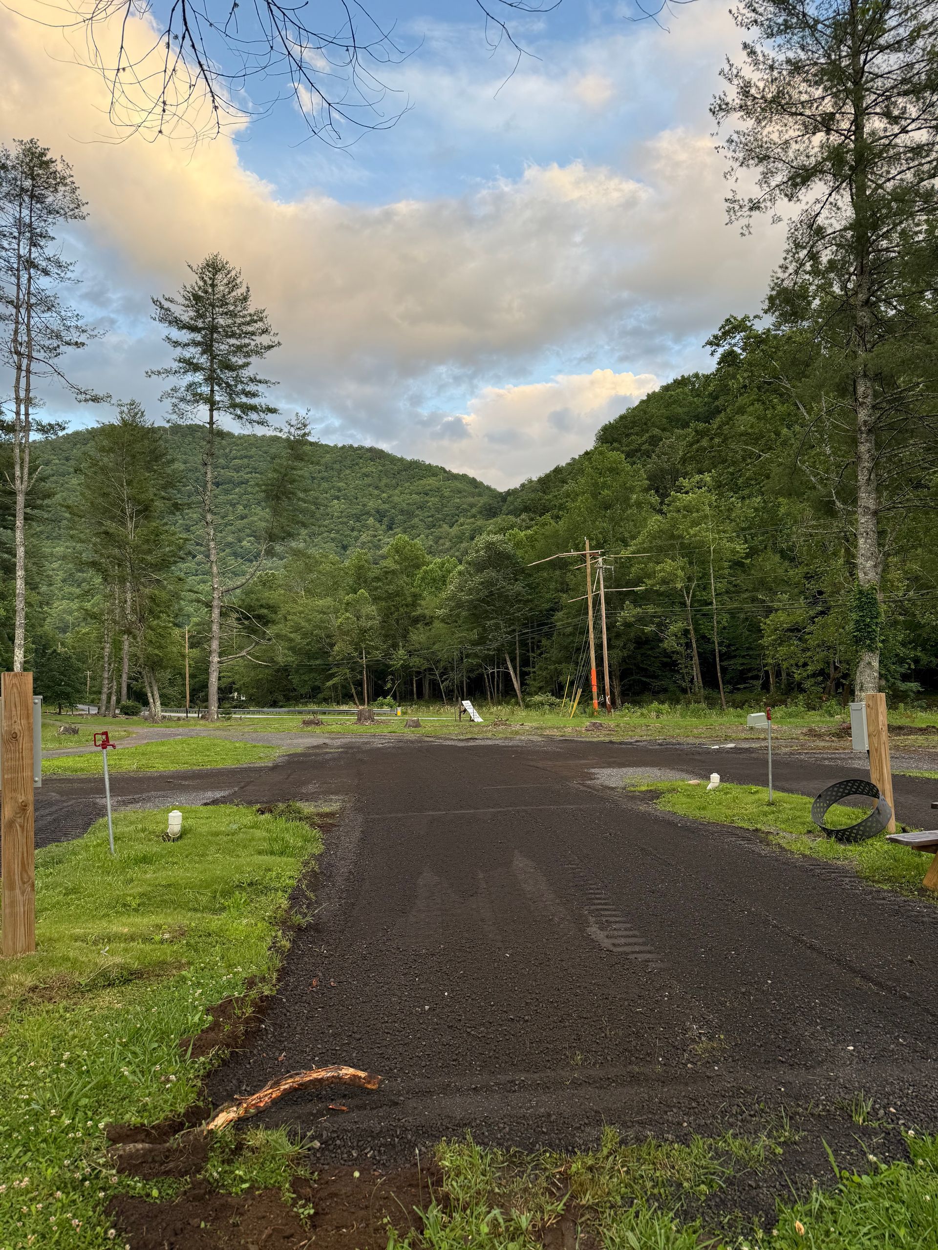Gravel path leads toward a forested valley under a cloudy sky. Green grass, tall trees, and power lines.