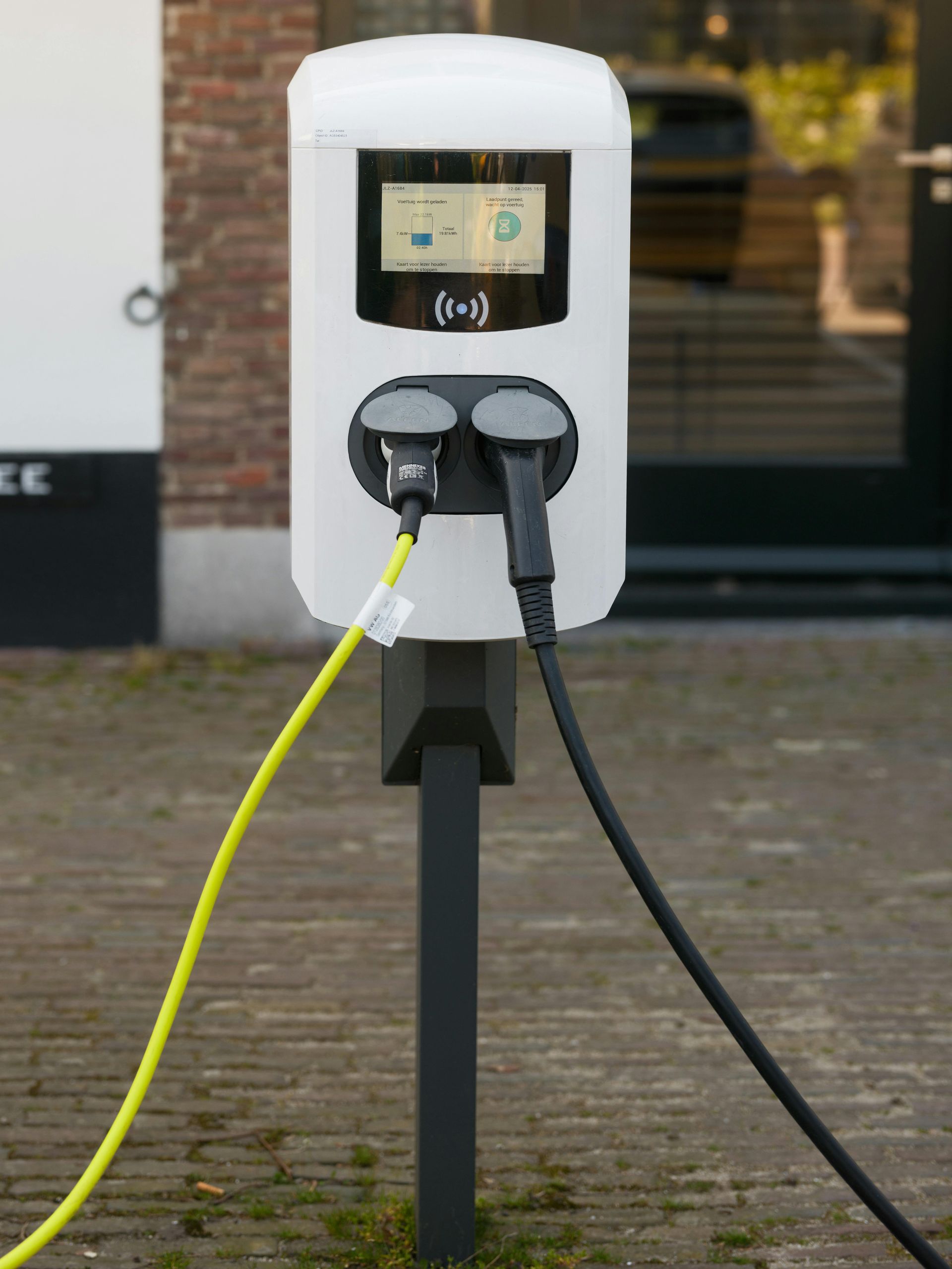 Borne de recharge pour véhicules électriques avec deux câbles branchés, en extérieur. Boîtier blanc sur poteau noir.