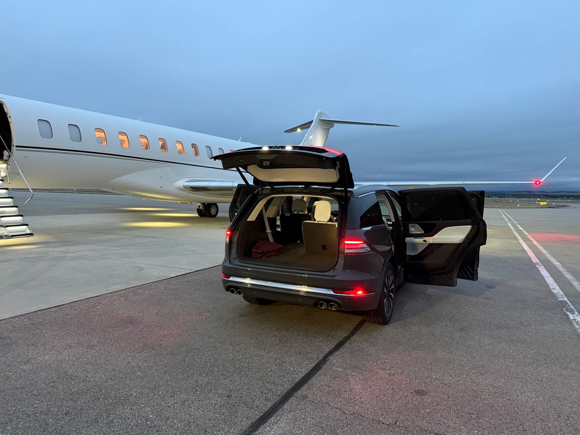 Black SUV on airport tarmac beside a private jet, rear hatch and door open at dusk