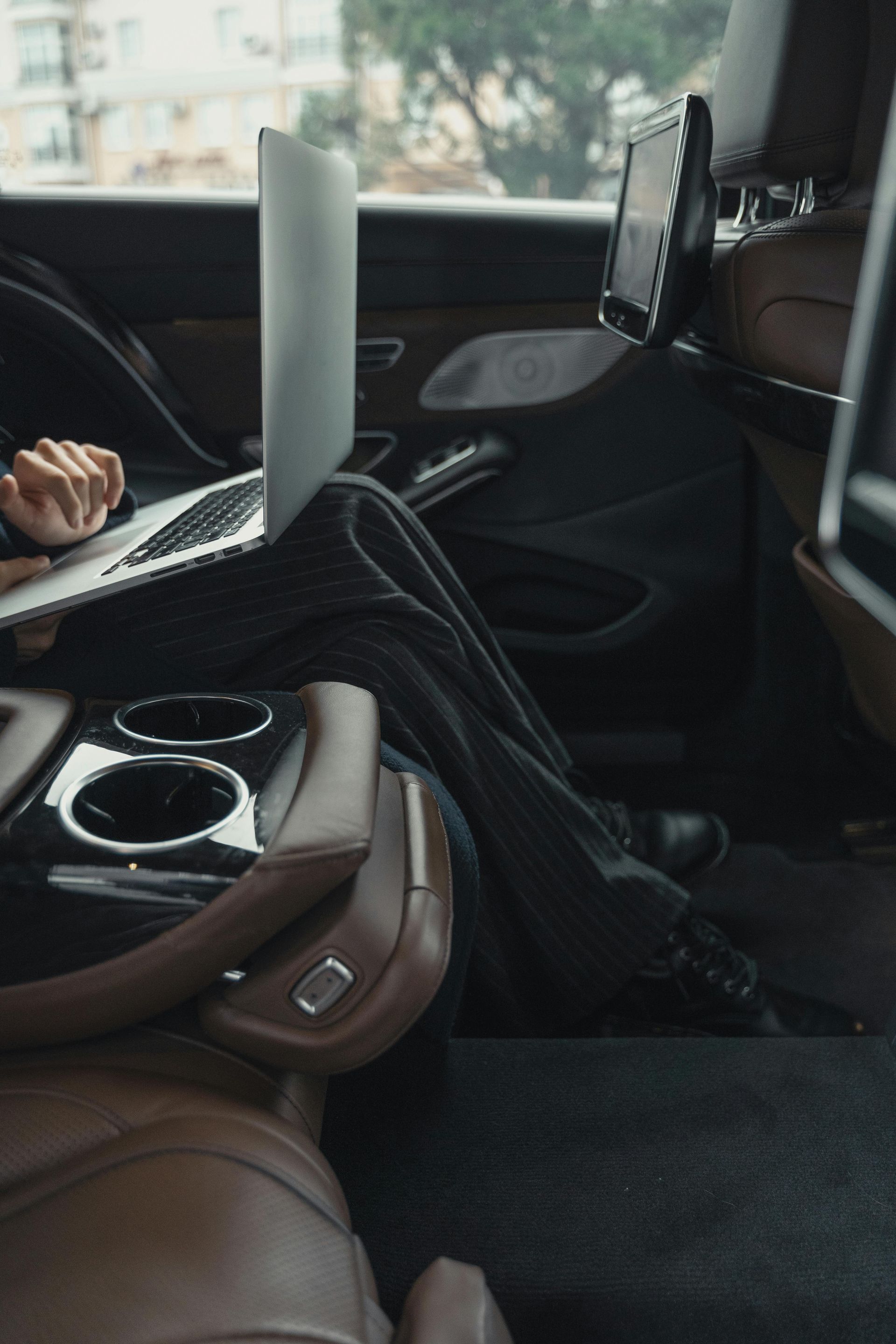 Person working on laptop inside a car, brown leather interior, two cup holders, purse.