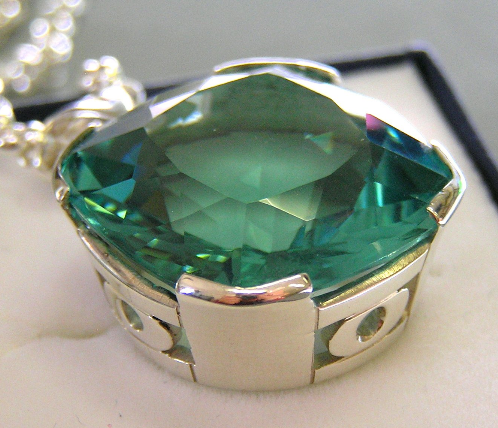 Primo piano di un ciondolo con una gemma sfaccettata color verde acqua, incastonata in una cornice decorativa d'argento su sfondo bianco.