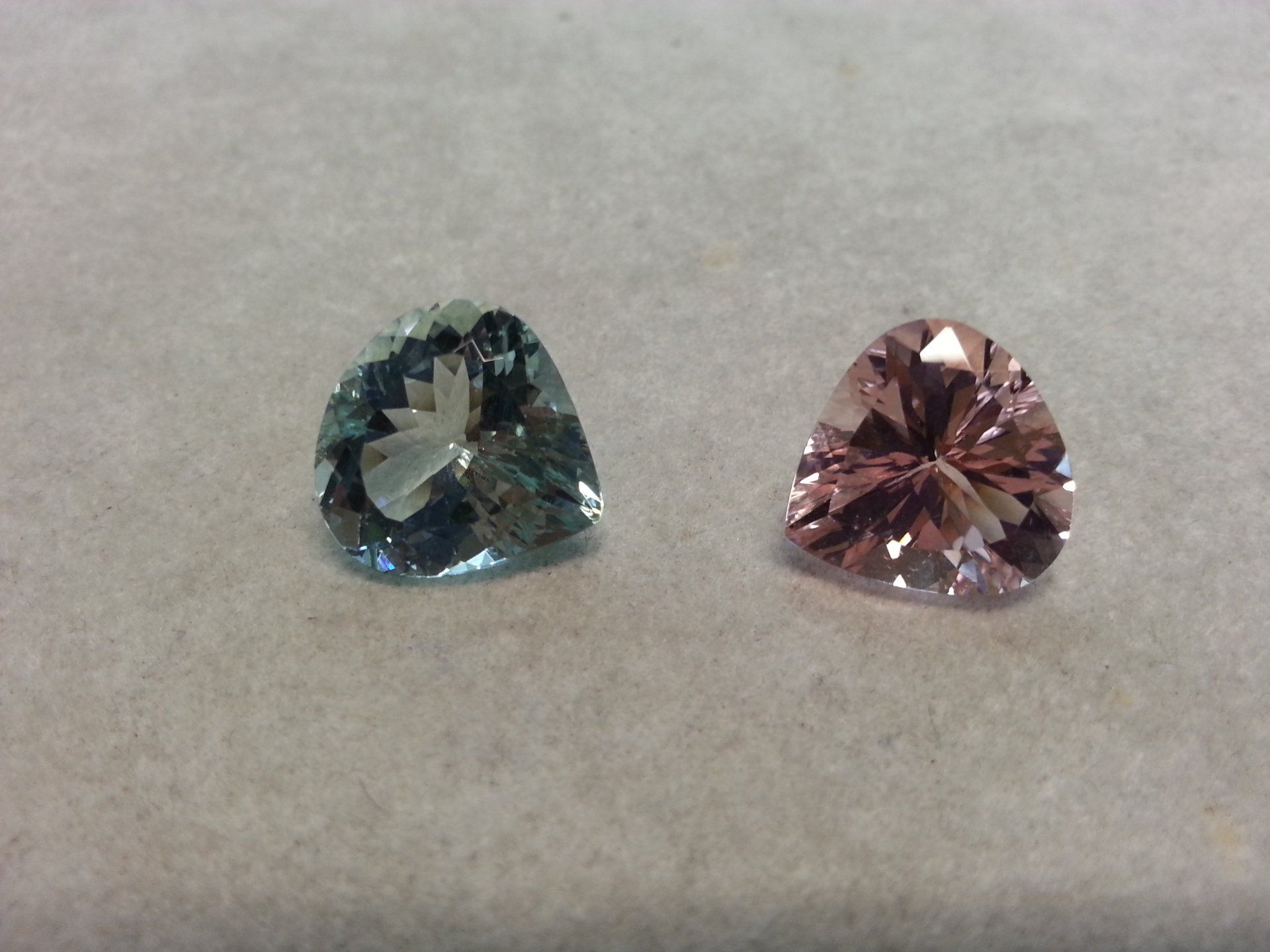 Due gemme triangolari sfaccettate, una blu-verde e una rosa chiaro, poggiate su una superficie piatta di colore grigio neutro.