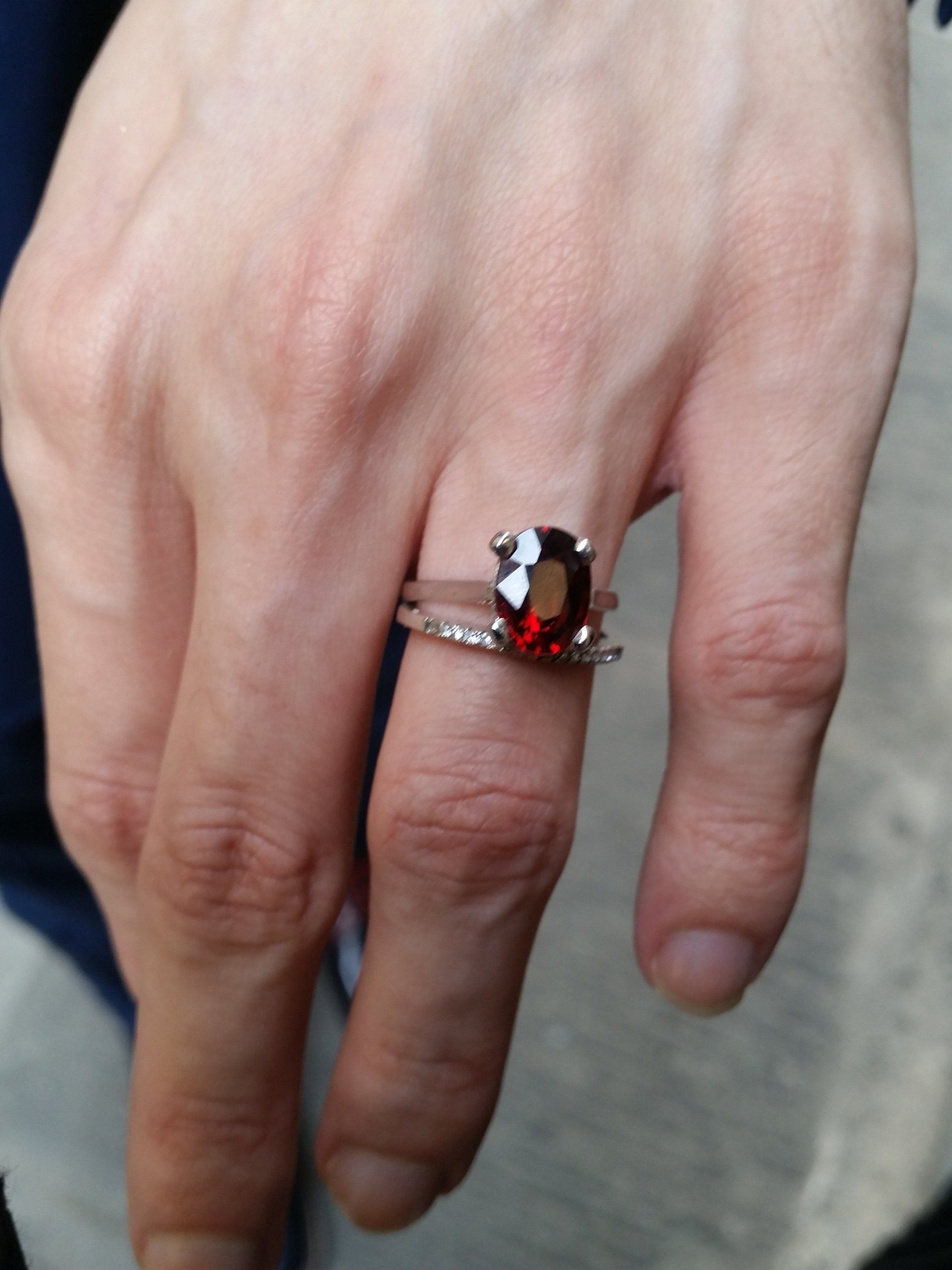 Una mano che indossa un anello d'argento con una vistosa gemma ovale di colore rosso intenso, sovrapposto a una sottile fascia tempestata di diamanti.