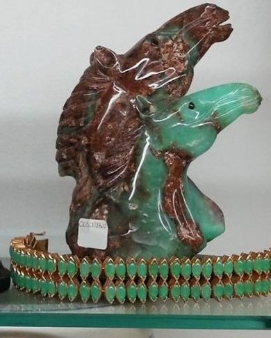 Una scultura di cavallo intagliata in pietre preziose bicolore marrone e verde, posta dietro un bracciale a doppia fila di pietre preziose verdi.