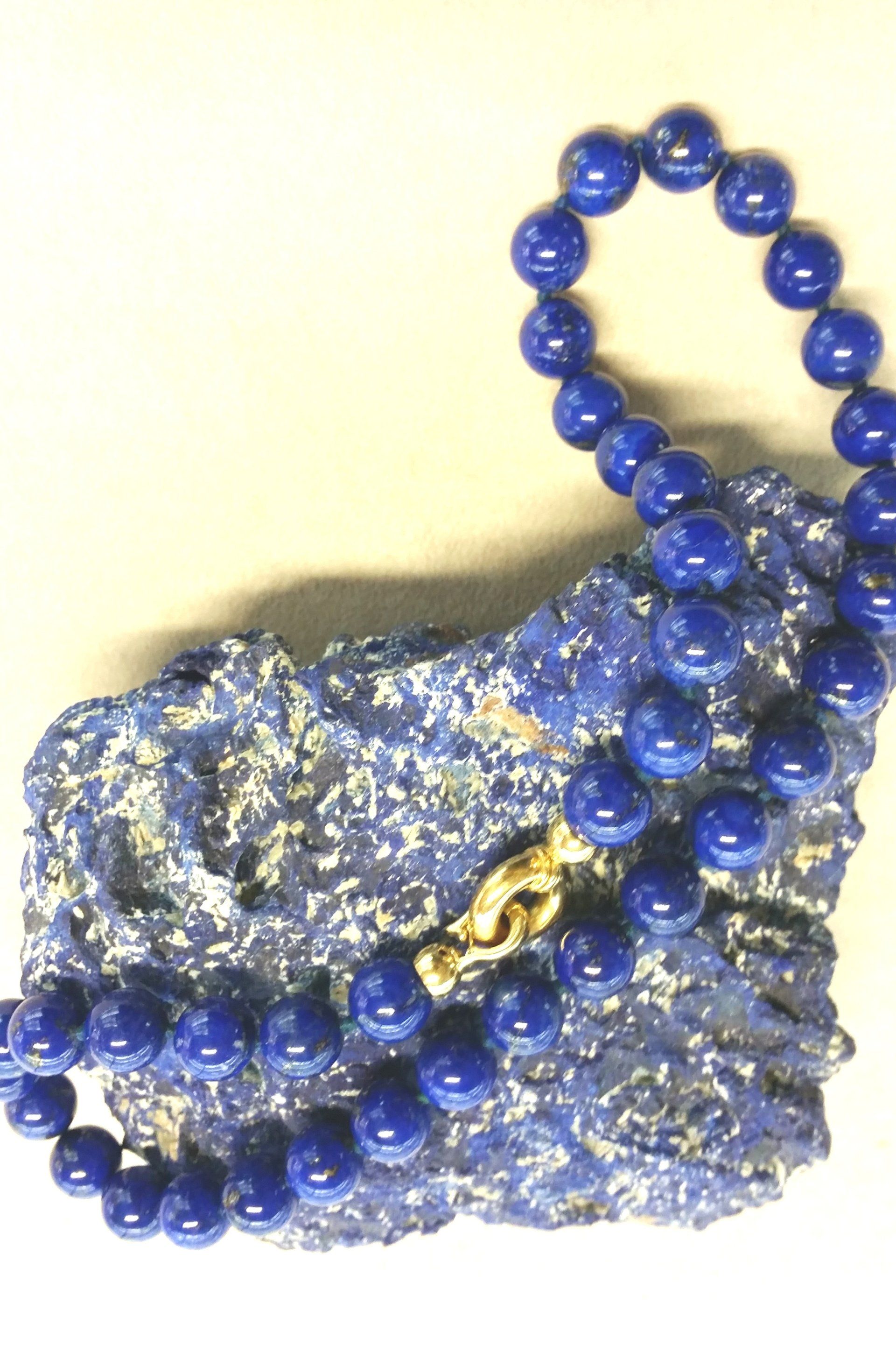 Una collana di vivaci perle di lapislazzuli blu con una chiusura in oro, poggiata su un pezzo grezzo e materico dello stesso minerale.