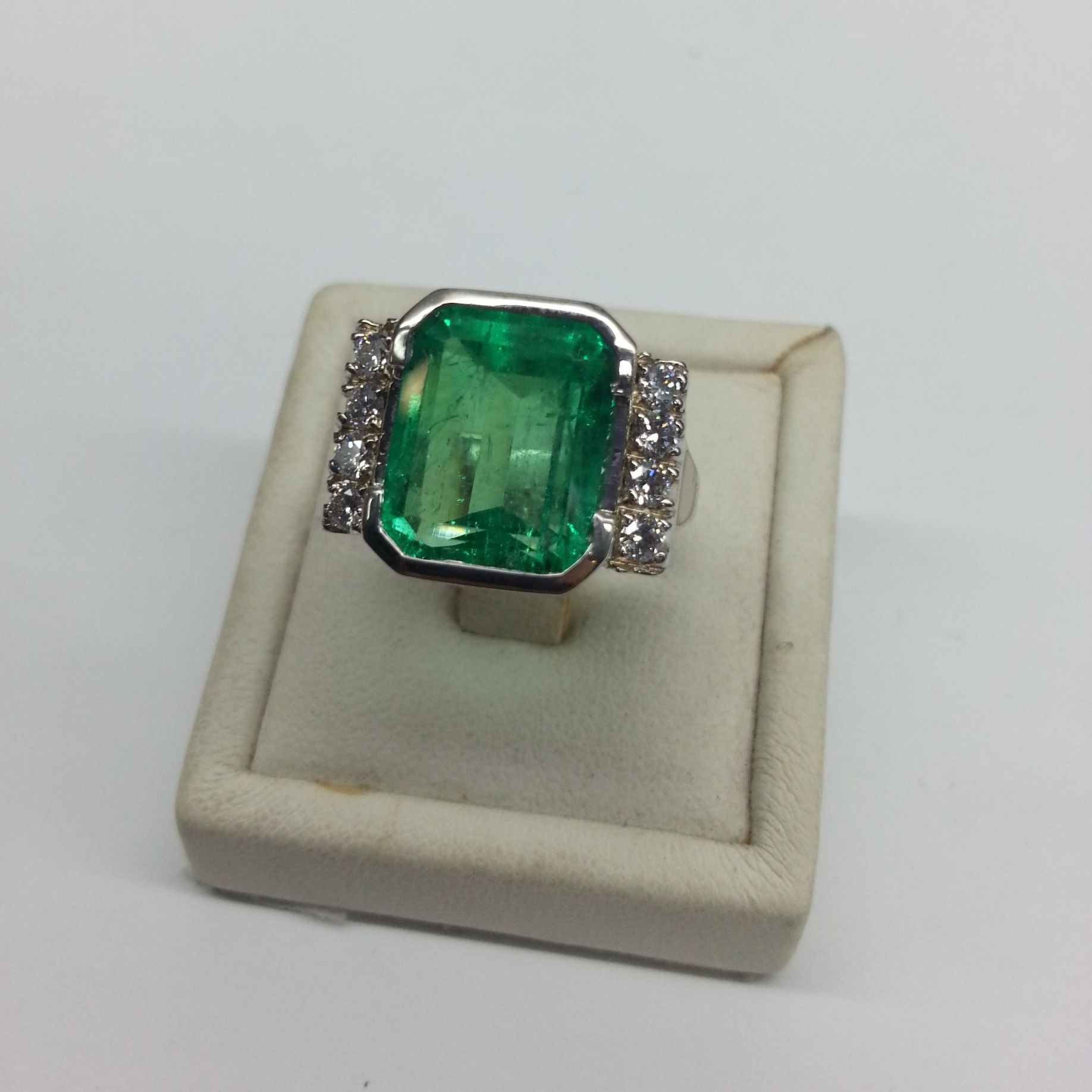 Un grande anello con una gemma verde taglio smeraldo, affiancata da file di piccoli diamanti bianchi, esposto su un cuscino portagioie color crema.