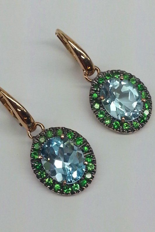 Un paio di orecchini pendenti ovali con una pietra centrale azzurra circondata da piccole gemme verdi incastonate in una montatura in oro rosa.