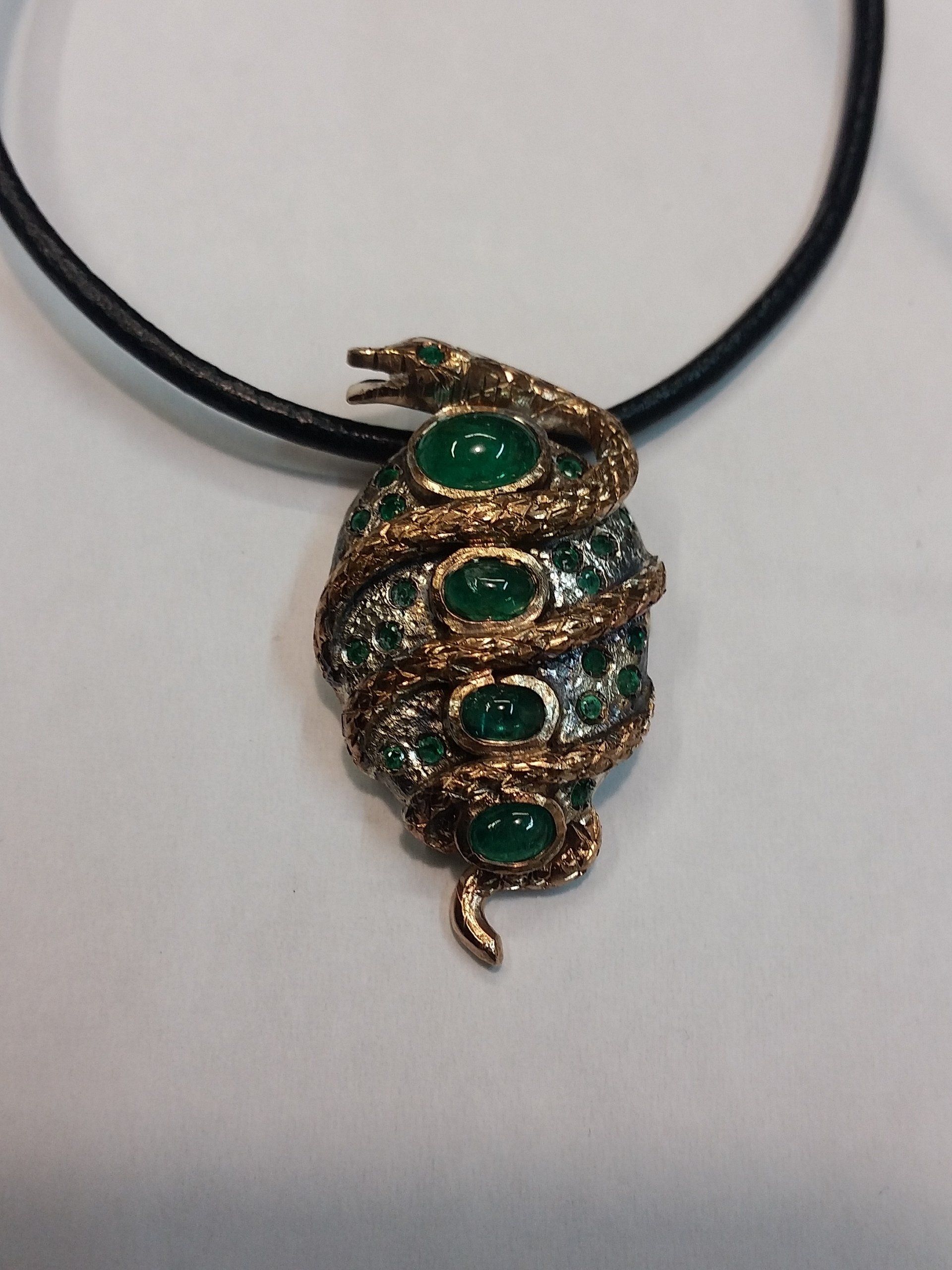 Un ciondolo a forma di serpente color oro, avvolto attorno a un gruppo di pietre verde scuro a taglio cabochon, appeso a un cordino nero.