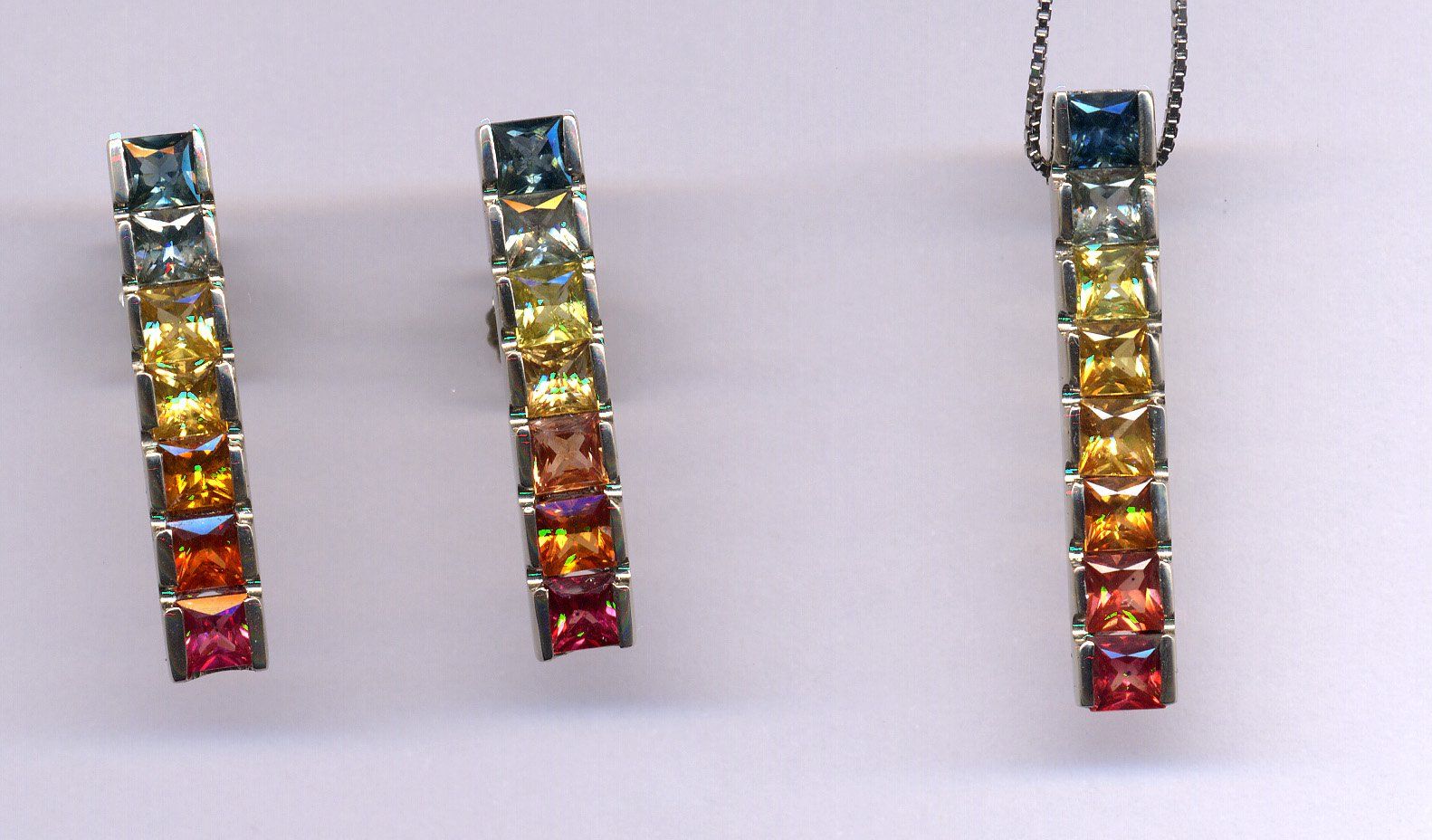 Un set di gioielli composto da due orecchini e un ciondolo abbinato, ciascuno con una sfumatura verticale arcobaleno di pietre preziose a taglio quadrato.