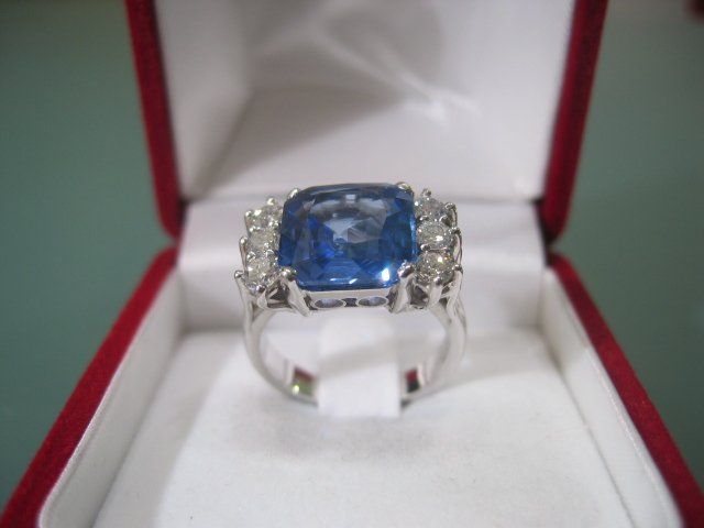 Un grande anello con gemma blu a taglio cuscino e dettagli in diamanti, esposto all'interno di un cofanetto portagioie aperto rosso e bianco.