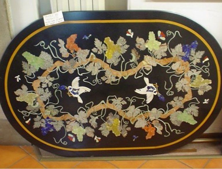 Un piano ovale da tavolo nero intarsiato con pietre colorate raffiguranti viti, foglie e due uccelli bianchi.
