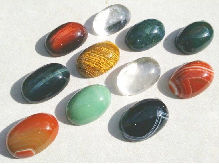 Dodici gemme ovali lisce e levigate, di vari colori tra cui verde, rosso, giallo, nero e trasparente, incastonate su una base bianca.
