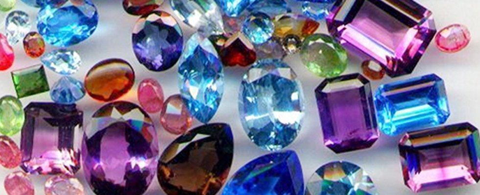 Una varietà di gemme colorate e sfaccettate di diverse forme, tra cui blu, viola e verdi, sparse su una superficie bianca.