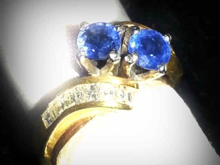 Anello in oro con due gemme blu rotonde e una fascia di pietre trasparenti più piccole incastonate su sfondo bianco.