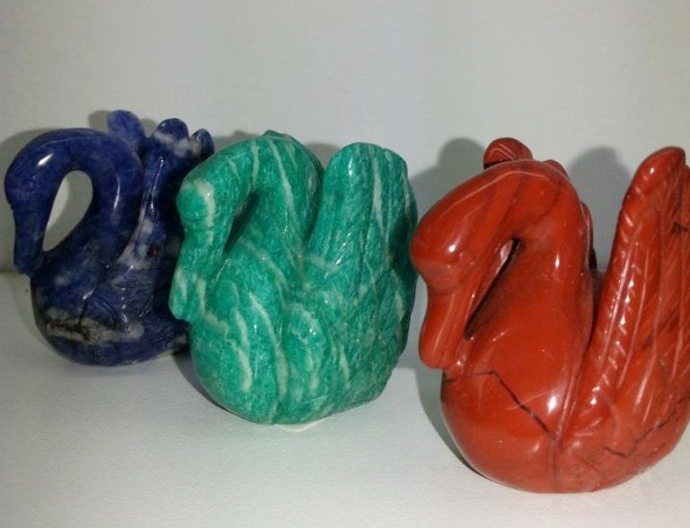 Tre statuette di cigni in pietra scolpita, nei colori blu, verde e rosso, sono allineate su una superficie bianca.