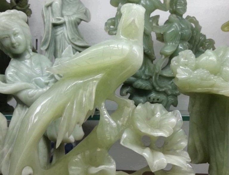 Statuetta in giada verde pallido raffigurante un uccello appollaiato tra fiori, esposta insieme ad altre sculture in pietra simili.