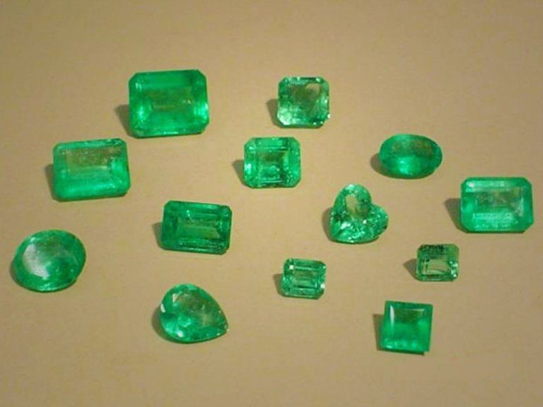 Una collezione di gemme di smeraldo verde sfaccettate, di varie forme e dimensioni, disposte su uno sfondo beige neutro.