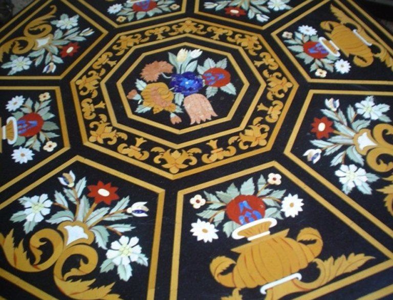 Un tavolo ottagonale in pietra nera con intricati intarsi floreali, un bouquet di fiori al centro e urne dorate.
