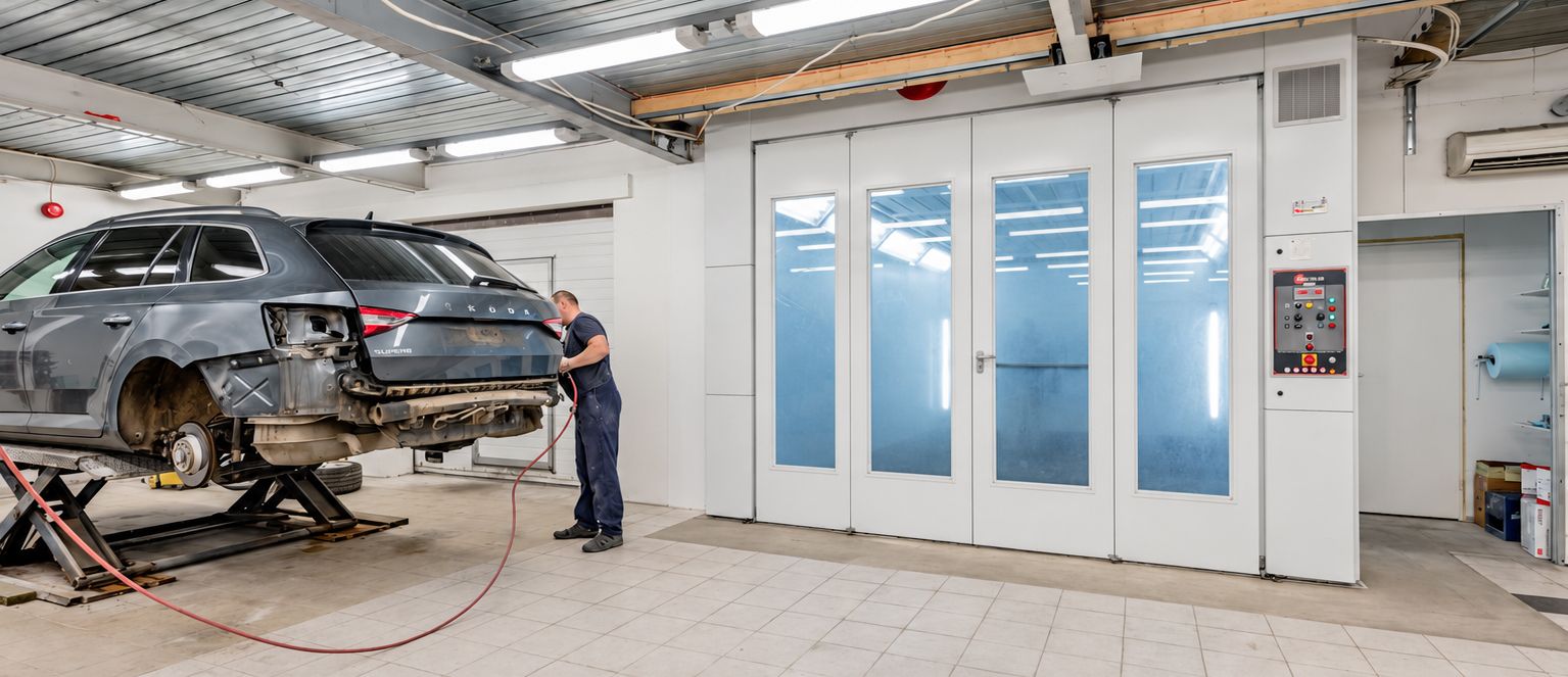 Car in an auto body shop being repaired beside a spray booth, with a mechanic working nearby.