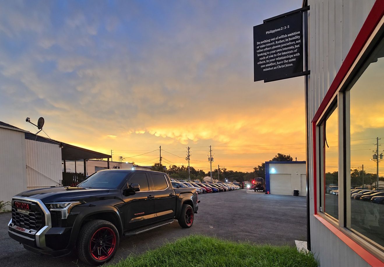 Black pickup truck at sunset beside a building, with a lit sign and reflective storefront windows.