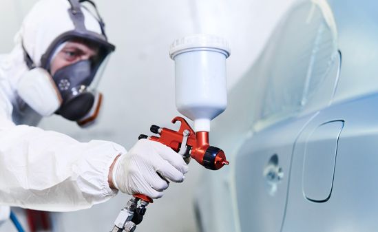 Worker in respirator spray-painting a light blue car in a body shop