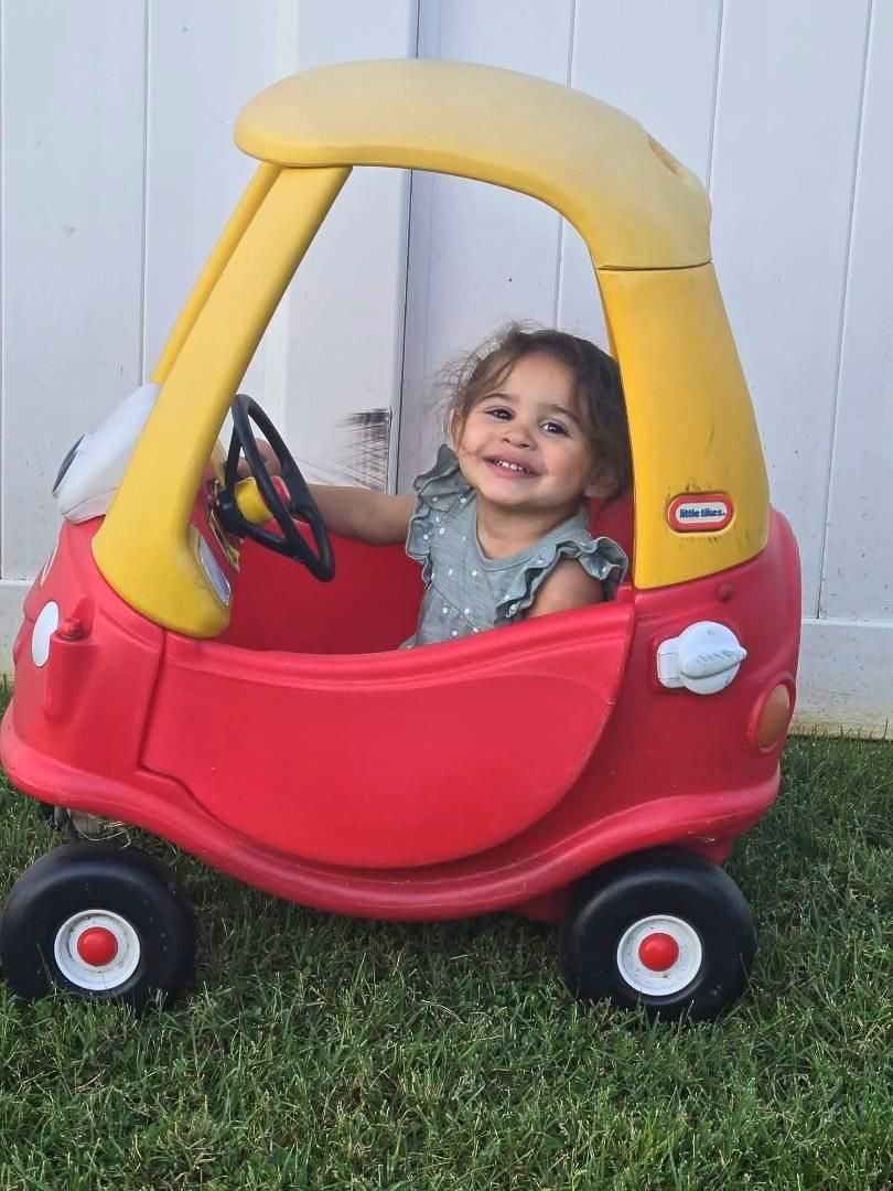 Smiling toddler in a red and yellow Little Tikes Cozy Coupe car on a grassy lawn.