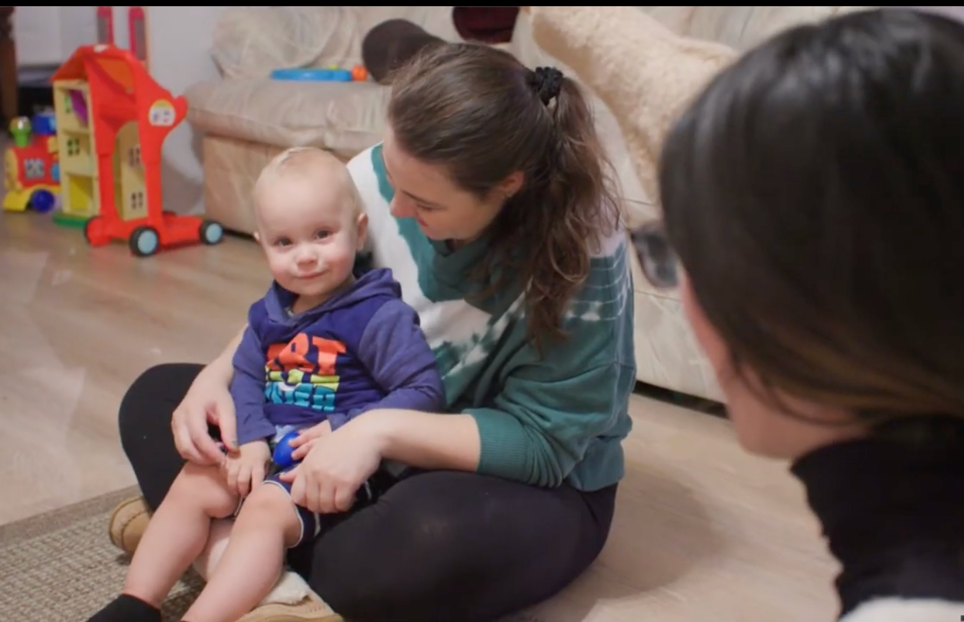 Woman with toddler on her lap, looking at another person. Living room setting with toys in background.