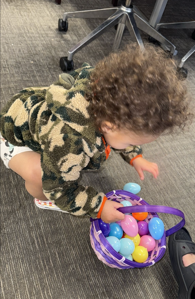 Child in camo jacket, squatting, reaching into purple Easter basket filled with colorful eggs on carpet.