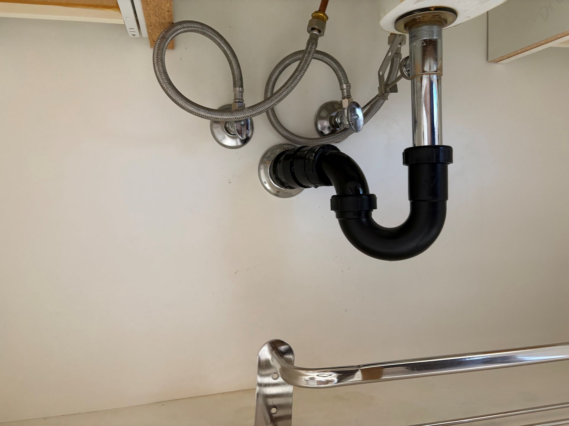 Under-sink plumbing with a black P-trap and chrome water lines against a white cabinet wall