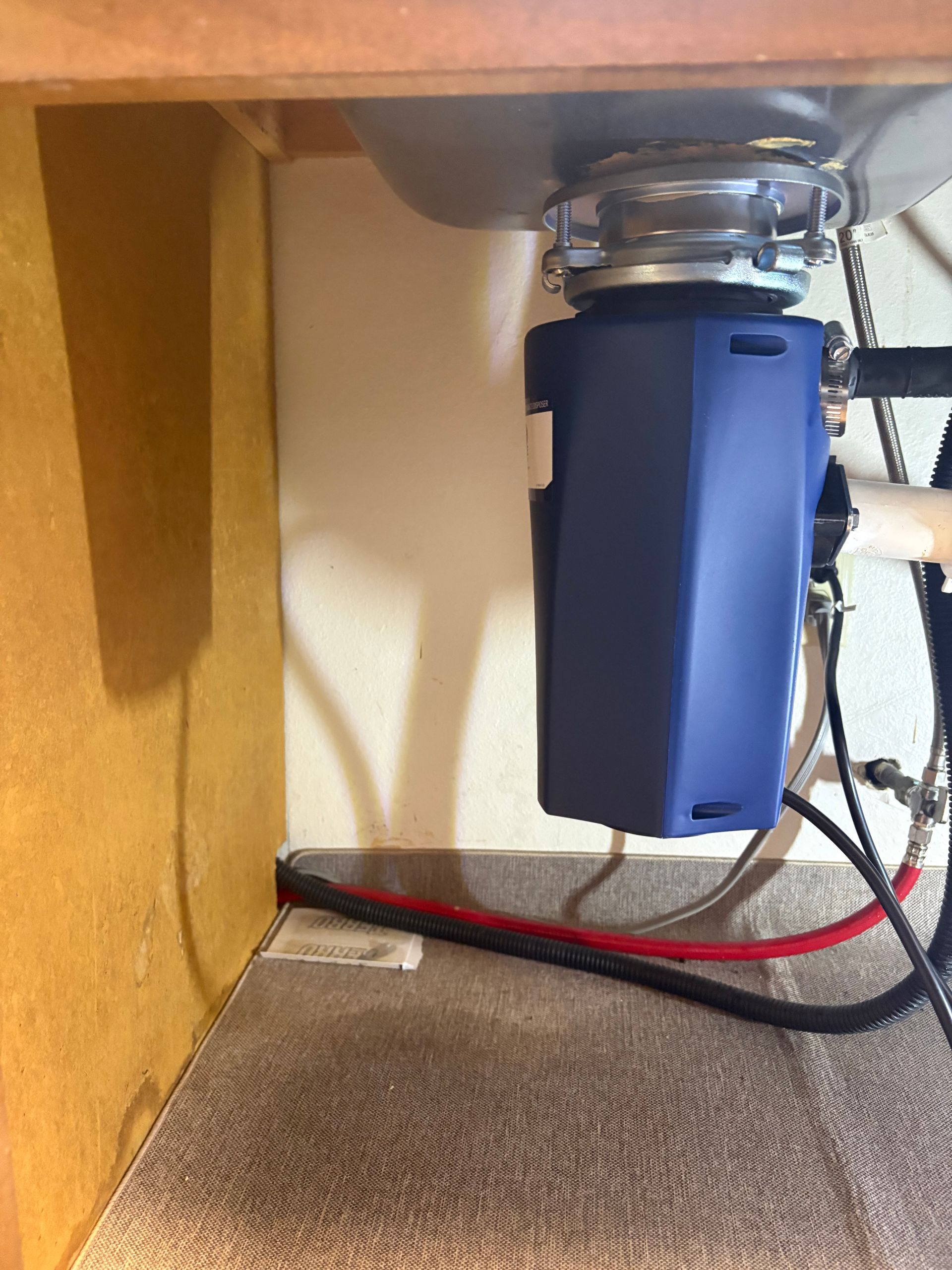 Under-sink garbage disposal with blue filter unit, hoses, and red wire on carpeted floor