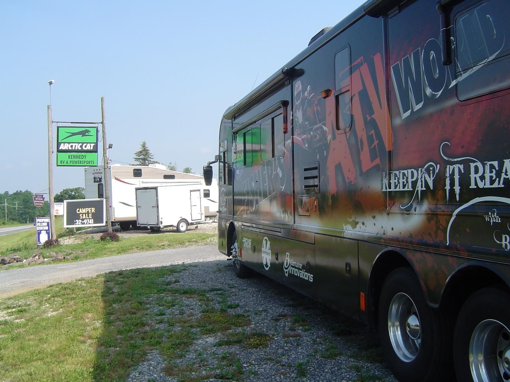 RV parked on gravel lot near an Arctic Cat sign and other campers on a sunny day.