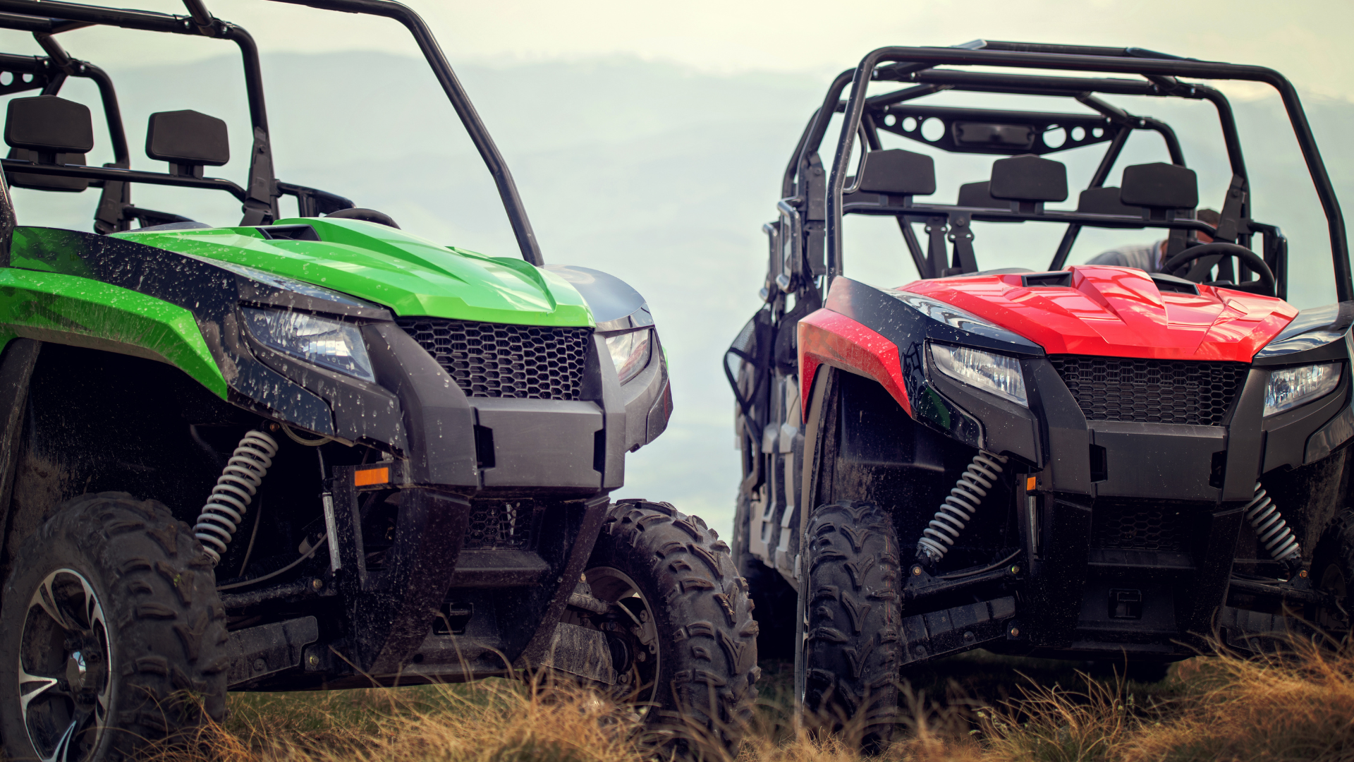 Green and red side-by-side UTVs parked on a grassy hill with a mountain backdrop.