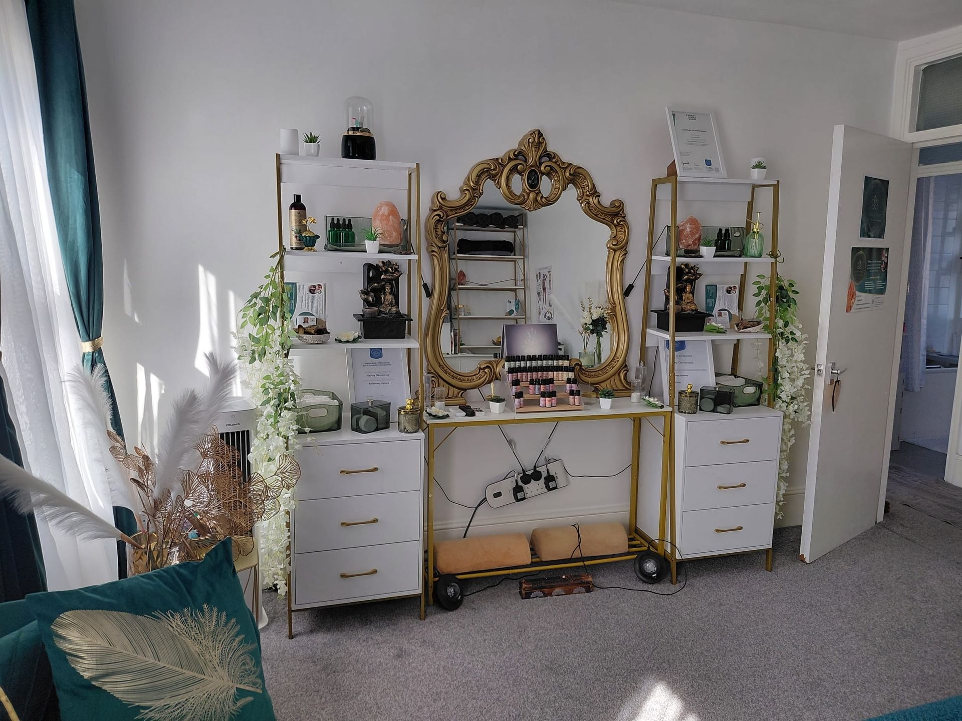 Bright bedroom vanity with ornate mirror, white drawers, plants, and teal chair by a window