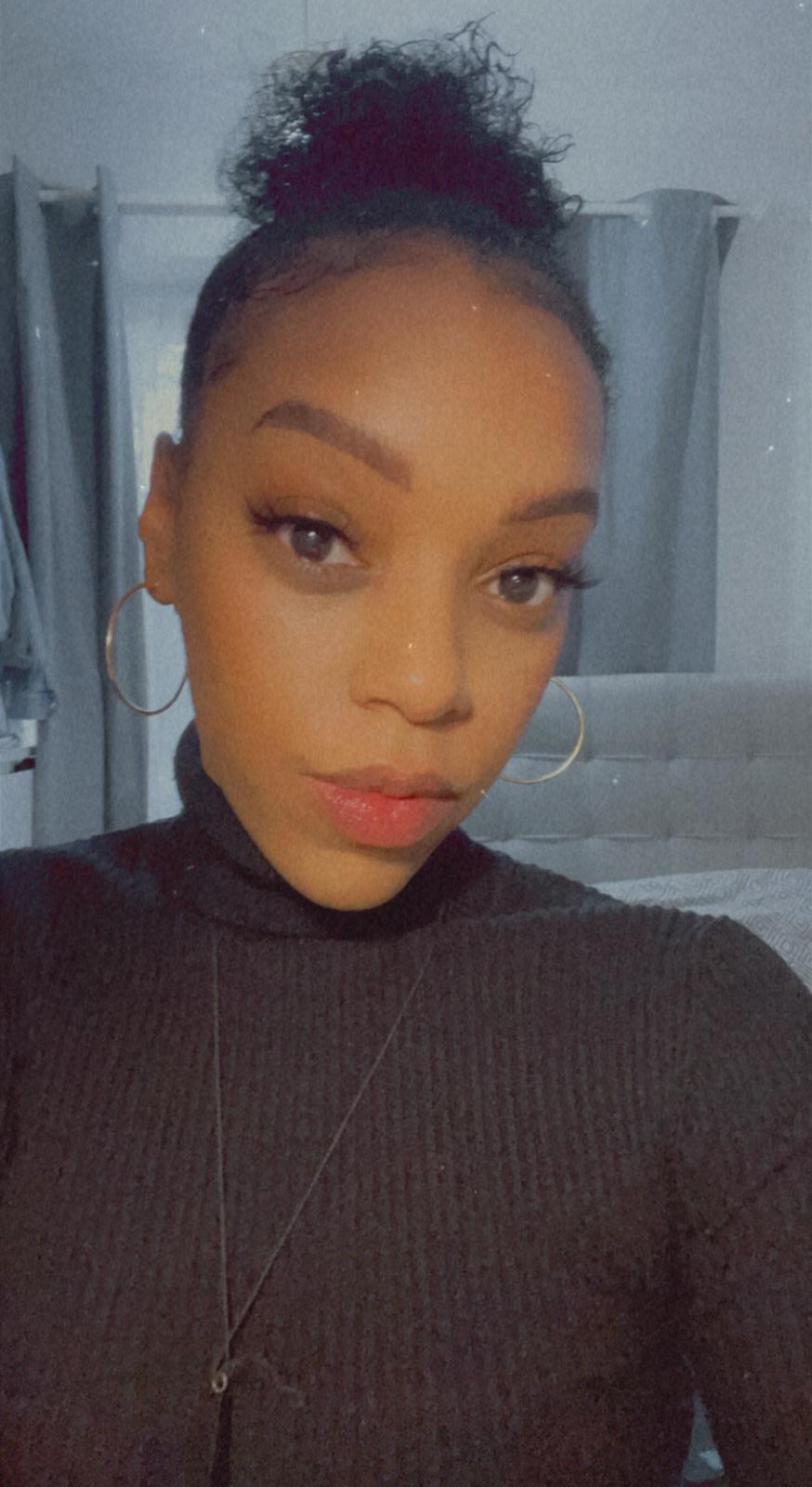 Portrait of a person in a black turtleneck, standing indoors with a calm expression and hair in a bun.