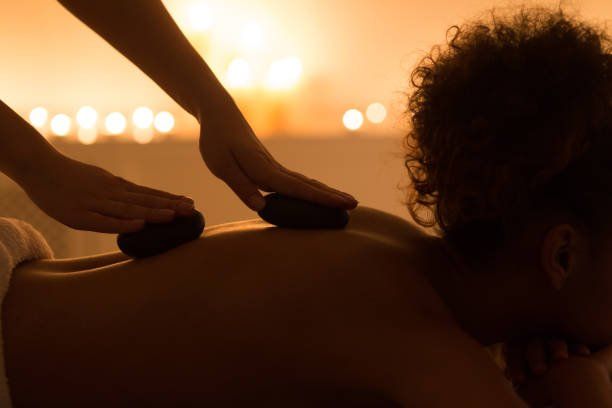 Massage therapist using hot stones on a person’s back in warm candlelit spa setting