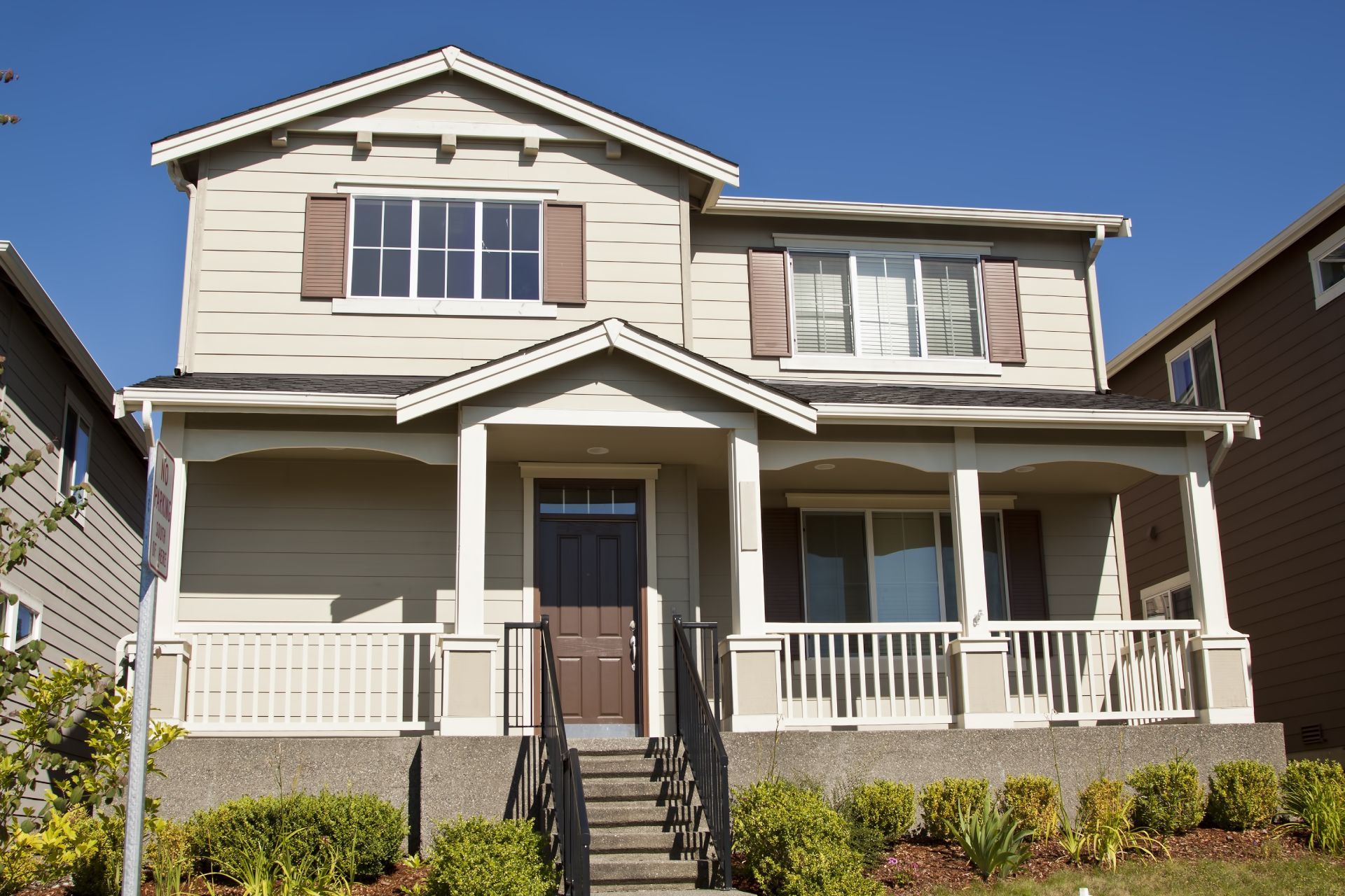 Two-story beige house with front porch, white trim, and brown shutters under a clear sky