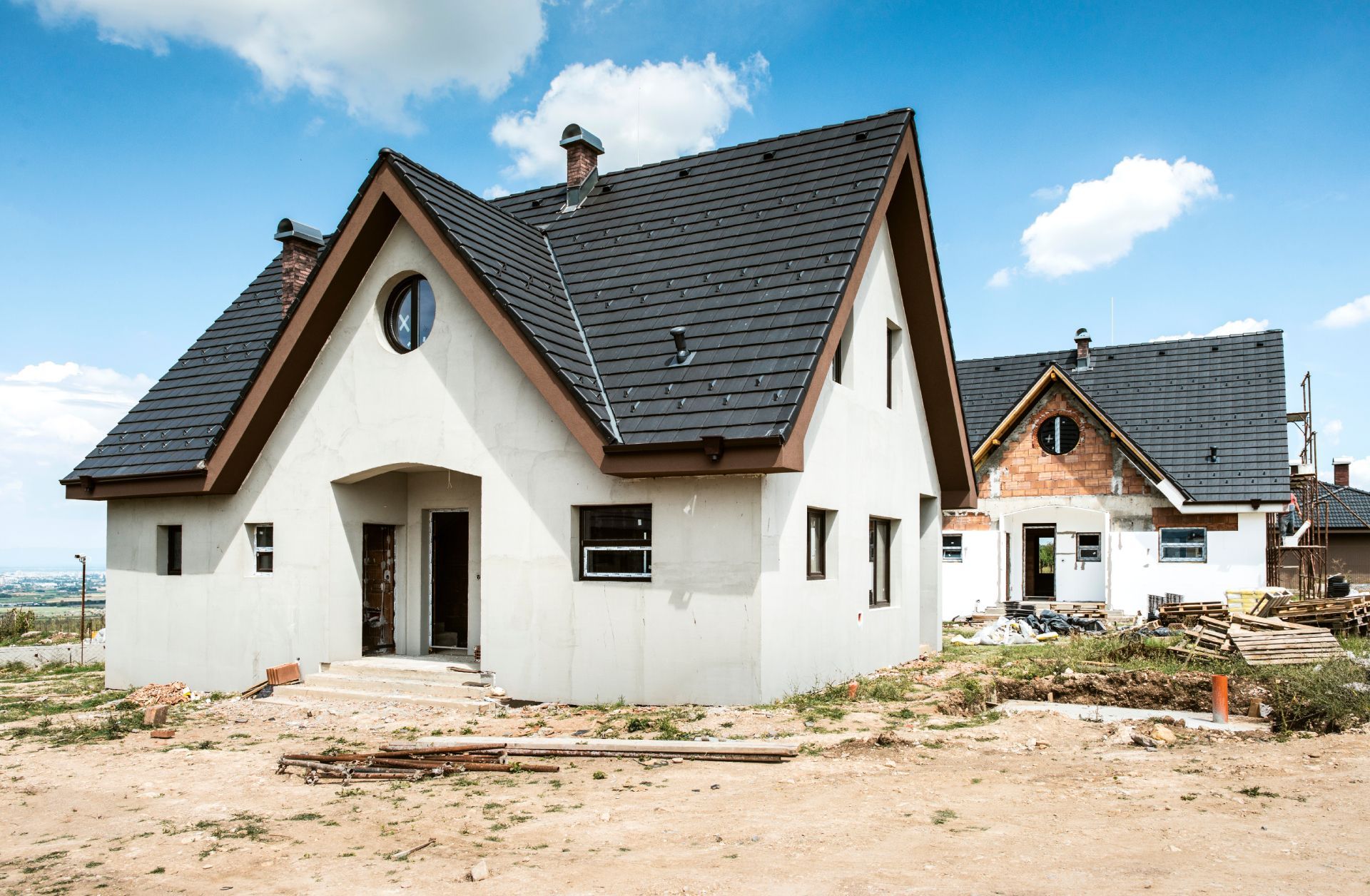Two unfinished white houses with dark roofs on a sandy construction site under a blue sky