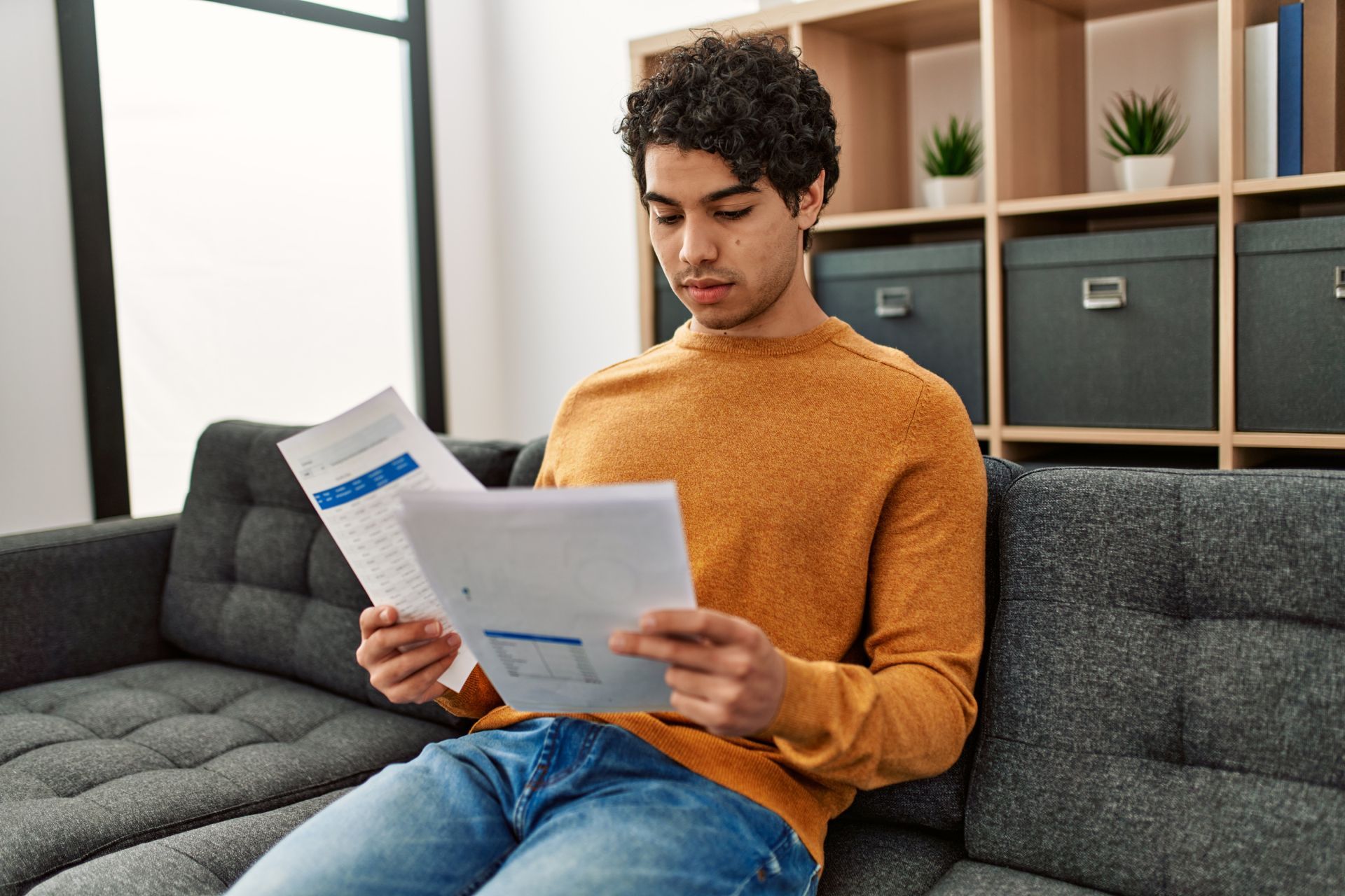 Person in an orange sweater reads papers on a gray couch in a bright living room.