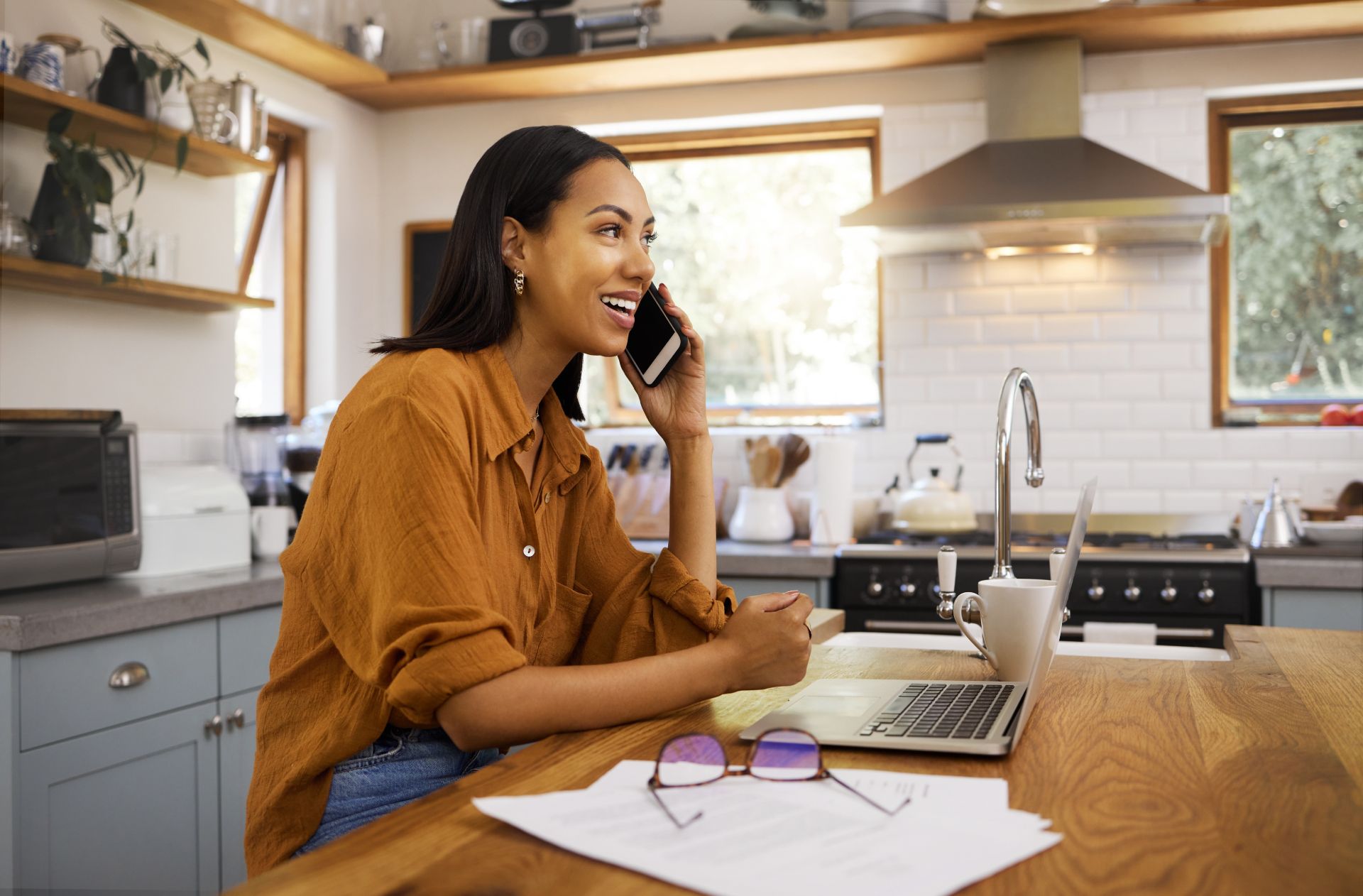Woman on a phone call in a bright kitchen, smiling beside a laptop and papers at a wooden table