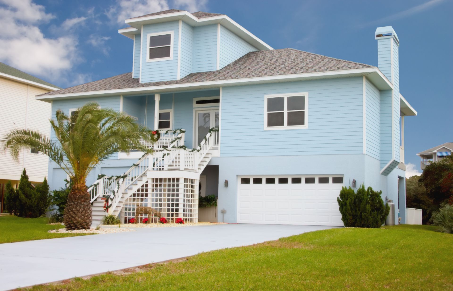 Light blue two-story house with white trim, front porch, garage, and palm tree on a green lawn.