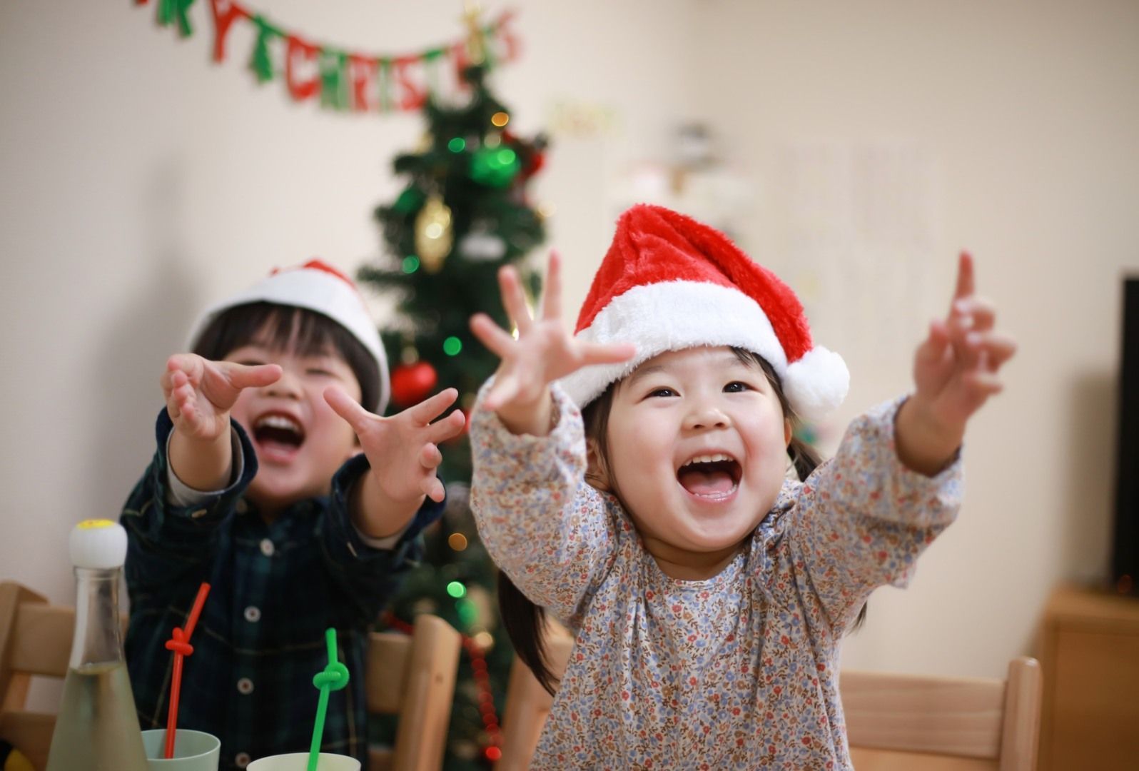 Two children in Santa hats excitedly reaching towards the viewer, Christmas tree in the background.