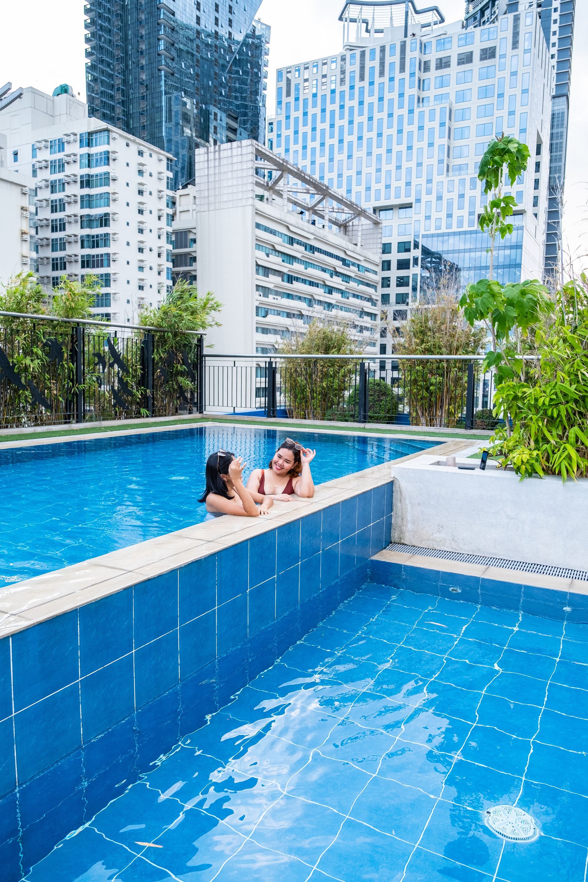 Two guests chatting in the rooftop pool of Y2 Residence Hotel Makati, surrounded by bamboo and city high-rises