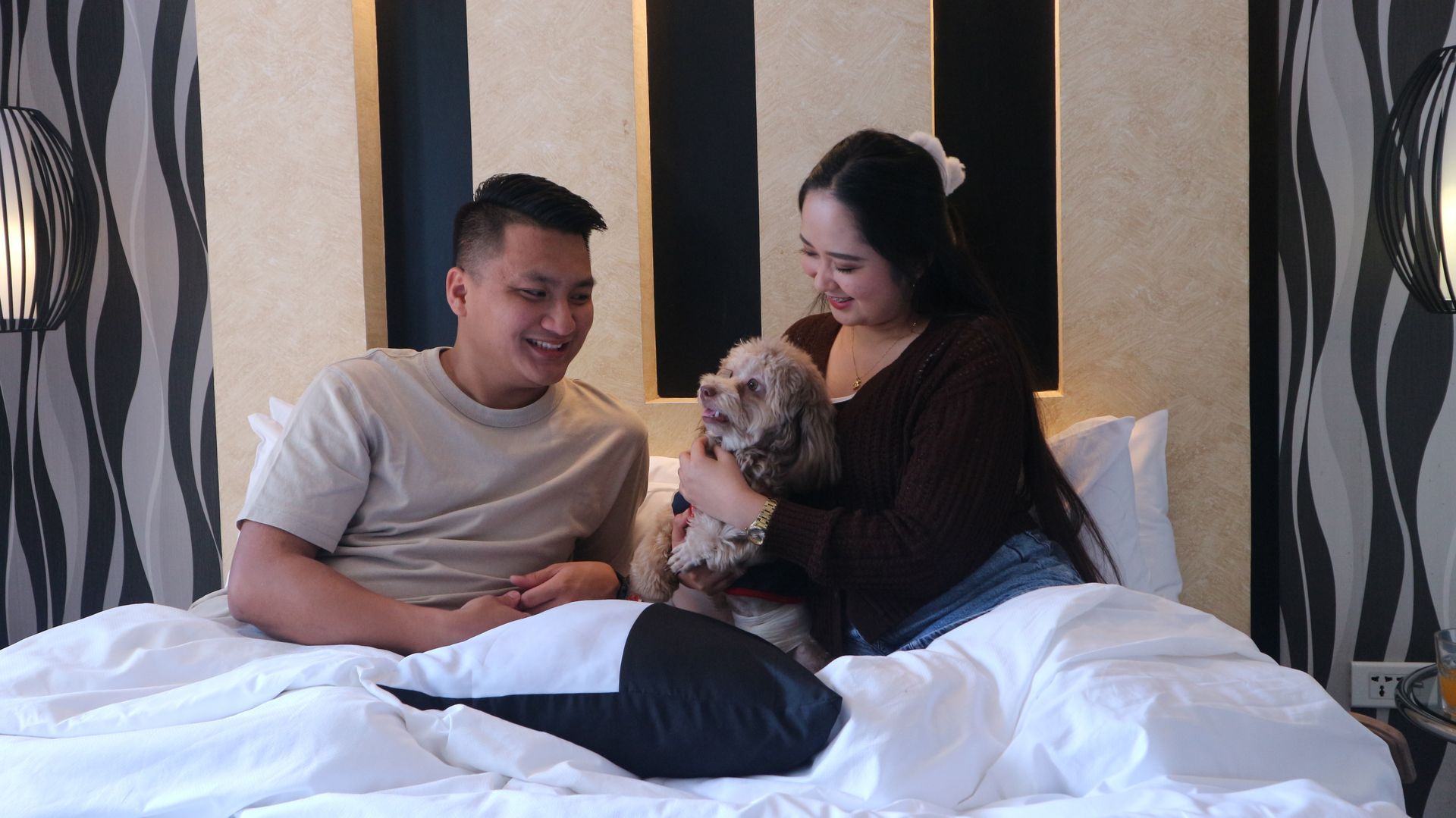 Couple cuddling their fluffy dog in bed in a guest room with a lit headboard at Y2 Residence Hotel Makati